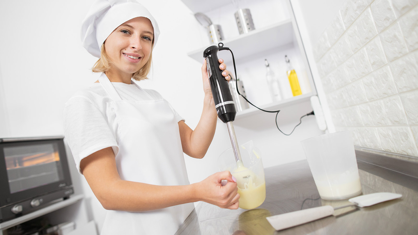 Young attractive girl pastry chef, wearing white apron and hat, prepares cream sauce or ganache for cake or cookies, holding a hand blender in her hand and mixes ingredients