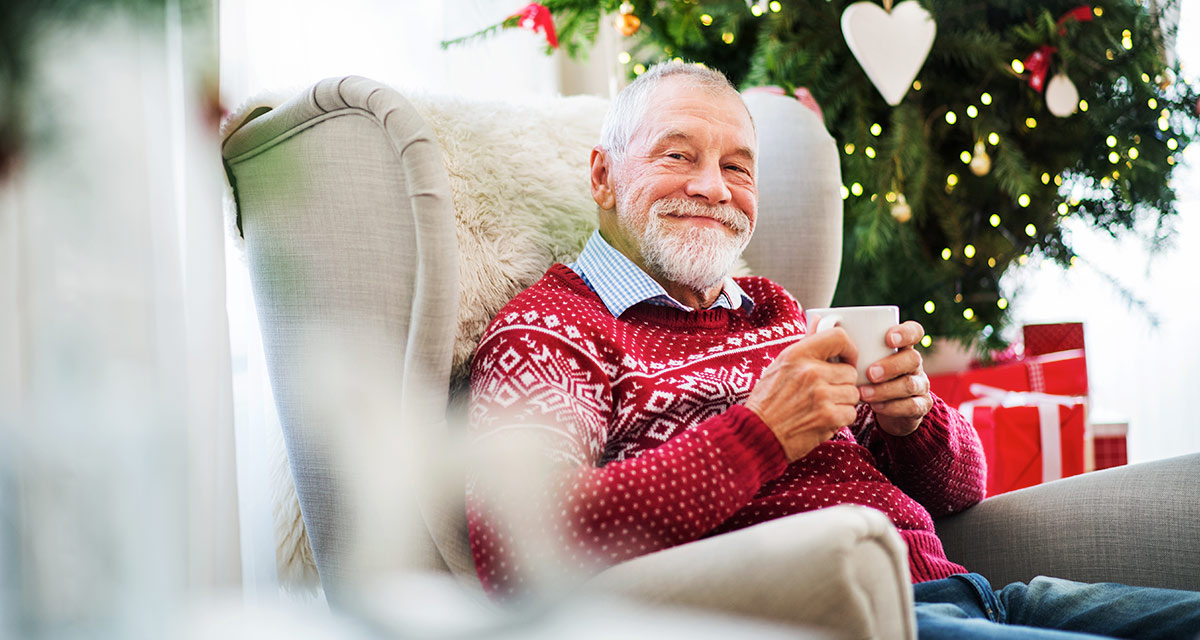Older man in red christmas sweater, smiling and sitting on chair drinking eggnog at Christmas.